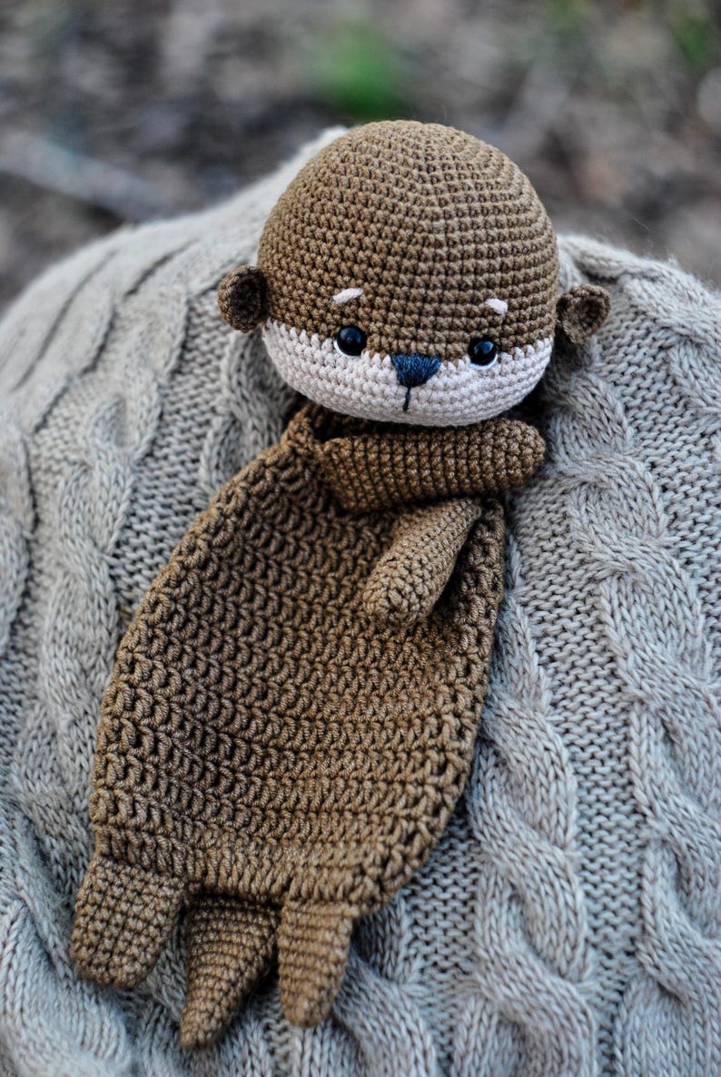 May include: A handmade crochet otter plush toy in shades of brown and cream. The otter has a detailed, textured design with small ears, a cream-colored face, and black eyes. The plush is lying on a gray cable-knit blanket.