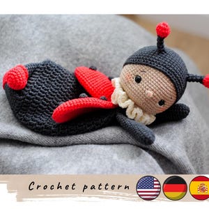 Può includere: Schema a uncinetto per una bambola coccinella con accenti neri e rossi. La bambola ha un viso dolce ed è sdraiata su una coperta grigia. Lo schema è disponibile in inglese, tedesco e spagnolo.
