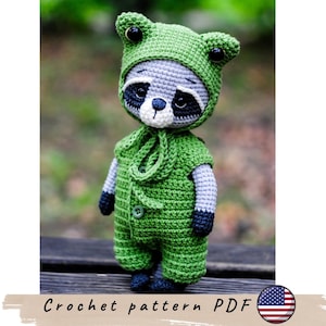 May include: A handmade crochet raccoon doll wearing a green outfit with a matching hat. The doll has a gray face with black markings around the eyes and a white muzzle. The text "Crochet pattern PDF" is at the bottom.