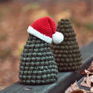 May include: Two crocheted Christmas trees, one wearing a red and white Santa hat, sitting on a wooden bench with fallen leaves.