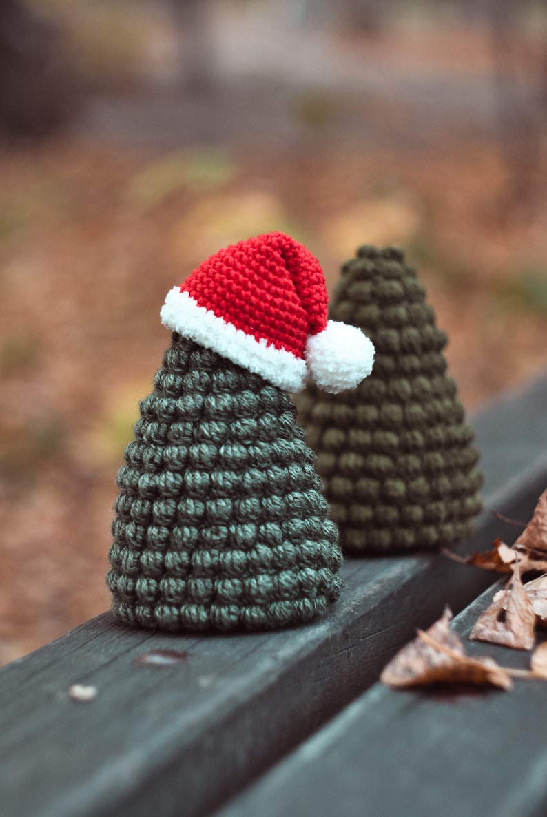 May include: Two crocheted Christmas trees, one wearing a red and white Santa hat, sitting on a wooden bench with fallen leaves.