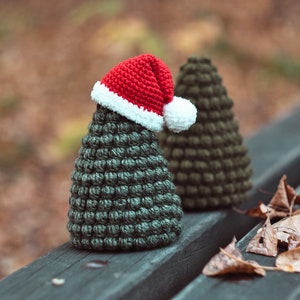 May include: Two green crocheted Christmas trees, one wearing a red and white Santa hat, sitting on a wooden bench with brown leaves scattered around them.