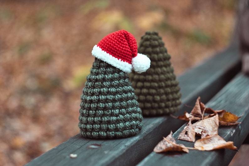 May include: Two green crocheted Christmas trees, one wearing a red and white Santa hat, sitting on a wooden bench with brown leaves scattered around them.