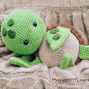 Pu&ograve; includere: Un giocattolo a forma di tartaruga fatto a mano all'uncinetto nei toni del verde, marrone e beige, appoggiato su una coperta grigia strutturata. La tartaruga presenta una testa verde con occhi neri e dettagli decorativi, un guscio marrone e pinne verdi.