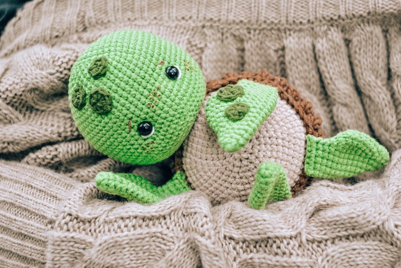Pu&ograve; includere: Un giocattolo a forma di tartaruga fatto a mano all'uncinetto nei toni del verde, marrone e beige, appoggiato su una coperta grigia strutturata. La tartaruga presenta una testa verde con occhi neri e dettagli decorativi, un guscio marrone e pinne verdi.