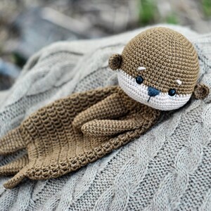 May include: A handmade crochet otter plush toy in shades of brown, white, and blue, resting on a gray cable-knit sweater. The otter has a detailed face with small black eyes and a blue nose. The plush is soft and cuddly.