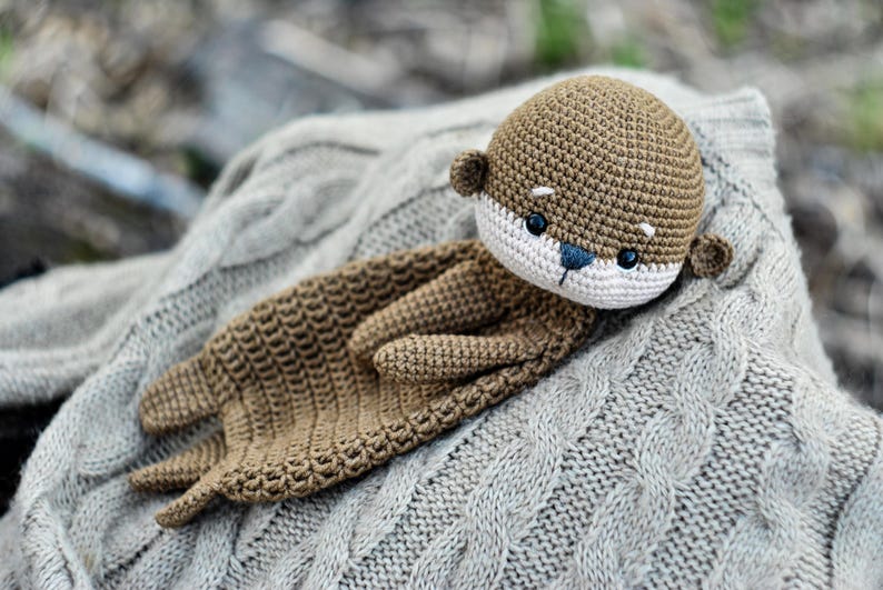 May include: A handmade crochet otter plush toy in shades of brown, white, and blue, resting on a gray cable-knit sweater. The otter has a detailed face with small black eyes and a blue nose. The plush is soft and cuddly.
