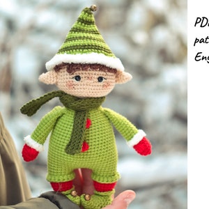 May include: A green crocheted elf doll with a white fur trim on the hat, red mittens, and a green scarf. The elf has a brown yarn hair and brown eyes. The doll is being held by a hand.