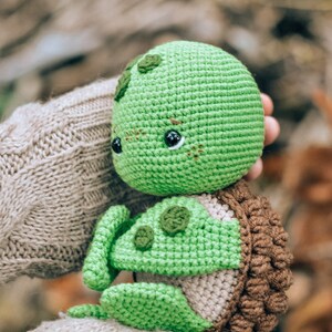 Pu&ograve; includere: Una tartaruga verde all'uncinetto fatta a mano con un guscio marrone e piccoli occhi neri. La tartaruga &egrave; tenuta tra le mani di una persona, con uno sfondo sfocato di foglie autunnali. La testa e gli arti della tartaruga sono di un verde vibrante.