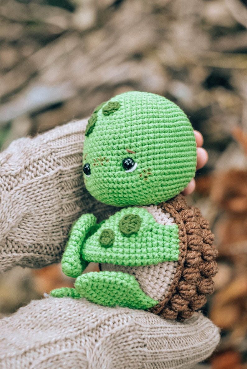 Pu&ograve; includere: Una tartaruga verde all'uncinetto fatta a mano con un guscio marrone e piccoli occhi neri. La tartaruga &egrave; tenuta tra le mani di una persona, con uno sfondo sfocato di foglie autunnali. La testa e gli arti della tartaruga sono di un verde vibrante.