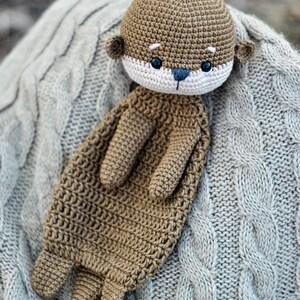 May include: A handmade crochet otter plush toy in shades of brown, beige, and blue. The otter has a detailed crocheted texture, with small ears, a white face, and blue eyes. The plush is resting on a gray cable-knit blanket.
