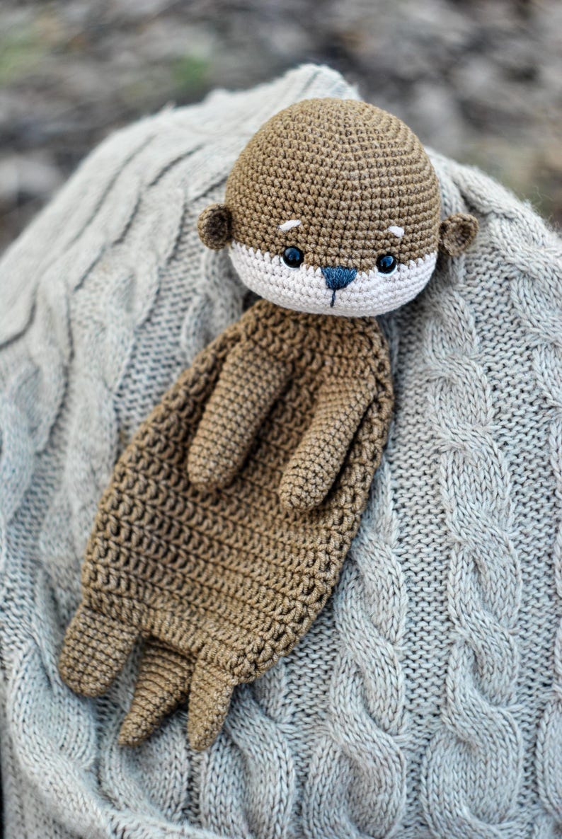 May include: A handmade crochet otter plush toy in shades of brown, beige, and blue. The otter has a detailed crocheted texture, with small ears, a white face, and blue eyes. The plush is resting on a gray cable-knit blanket.