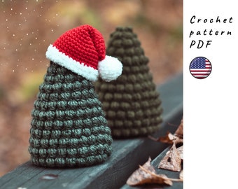 May include: Two crocheted Christmas trees, one wearing a red Santa hat with a white pom-pom. The trees are green and sitting on a wooden surface with fallen leaves.