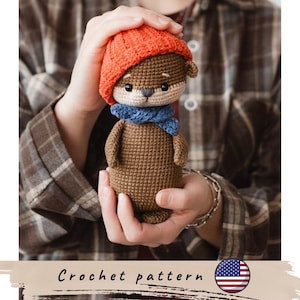 May include: A crocheted otter doll wearing an orange hat and blue scarf. The otter is brown with a beige face and small ears. The doll is held in a person's hands. The text "Crochet pattern" is at the bottom.