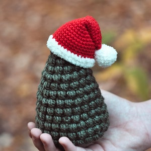 May include: A small crocheted Christmas tree with a red and white Santa hat. The tree is green and has a textured surface. The tree is being held in a person's hand.