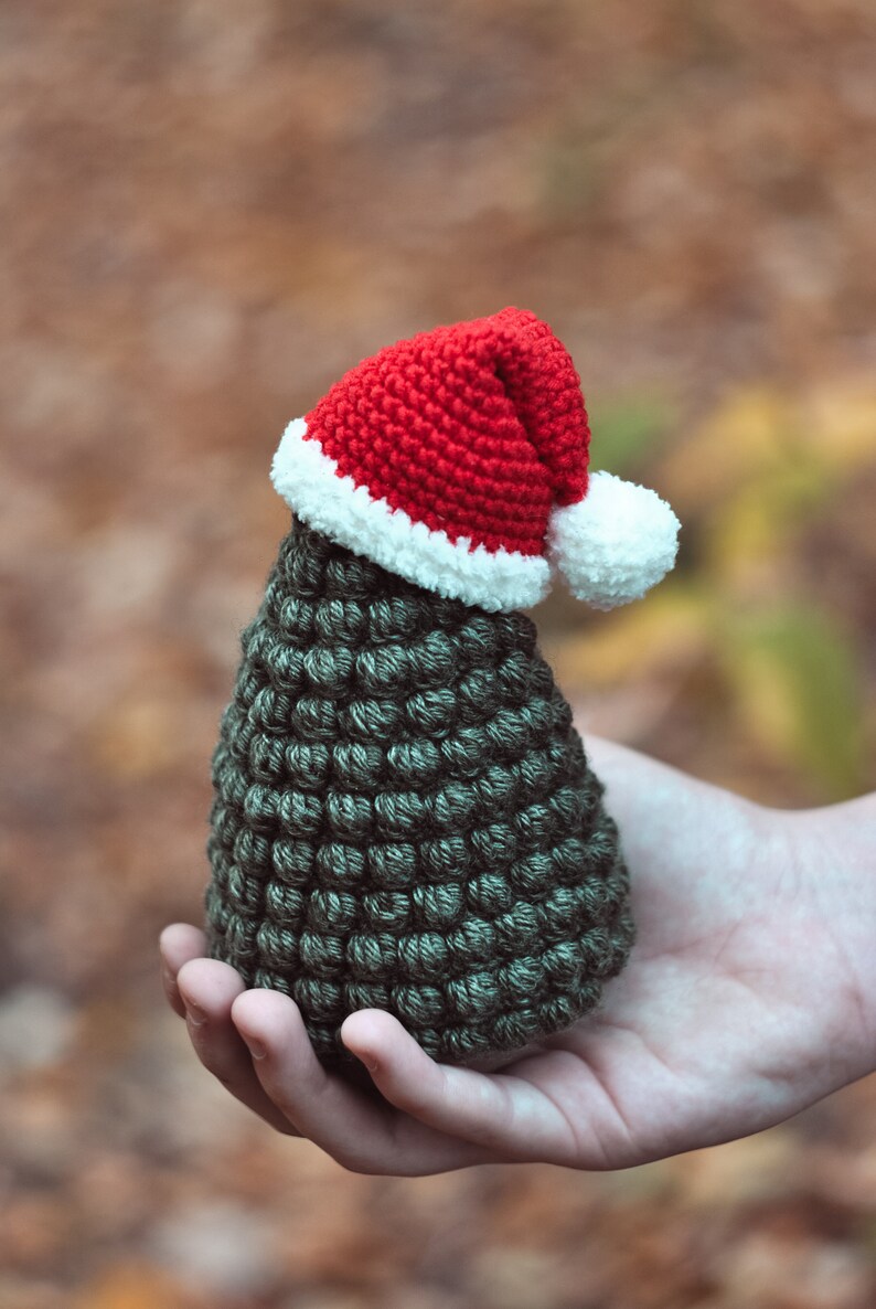 May include: A small crocheted Christmas tree with a red and white Santa hat. The tree is green and has a textured surface. The tree is being held in a person's hand.