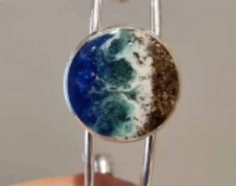 May include: A silver bangle bracelet with a round, blue, green, and brown resin inlay that resembles a beach scene.