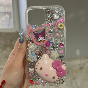 May include: Clear phone case with a pink Hello Kitty character, a black and white My Melody character, and various other charms and glitter.
