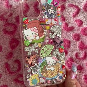 May include: Clear phone case decorated with Hello Kitty and other cartoon characters. The case features various charms, including a Hello Kitty popsicle, a rainbow, and a bowl of ramen. The background is a pink leopard print.