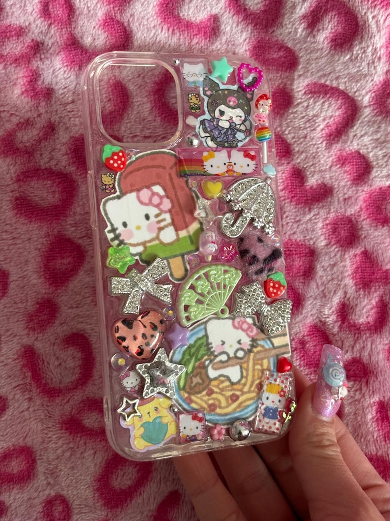 May include: Clear phone case decorated with Hello Kitty and other cartoon characters. The case features various charms, including a Hello Kitty popsicle, a rainbow, and a bowl of ramen. The background is a pink leopard print.