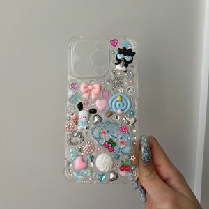 May include: Clear phone case with a variety of colorful 3D embellishments, including a cartoon dog, a bow, a star, and a swirl. The case is decorated with rhinestones and other small details.