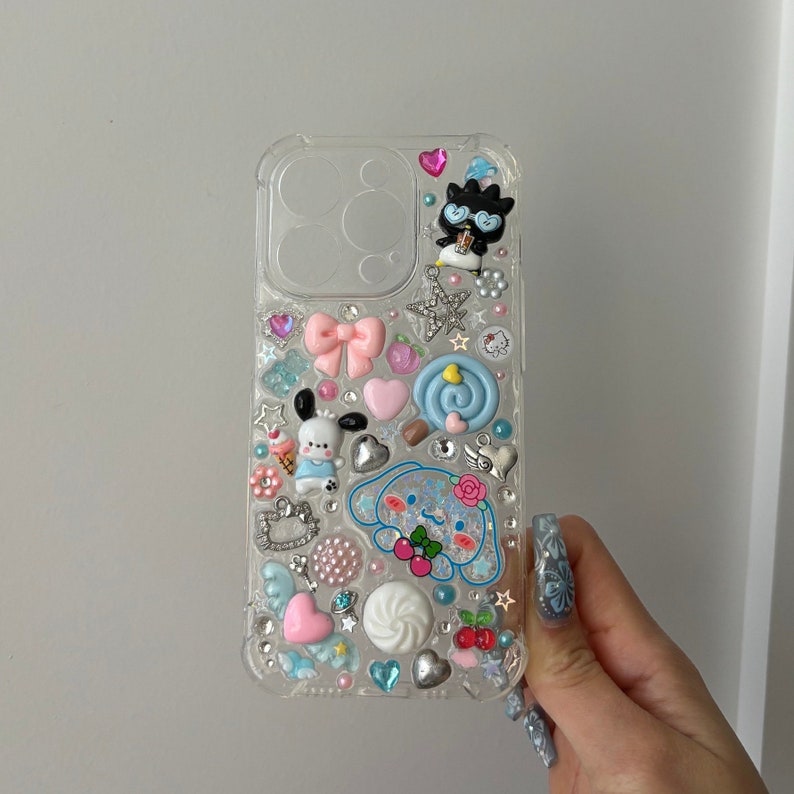 May include: Clear phone case with a variety of colorful 3D embellishments, including a cartoon dog, a bow, a star, and a swirl. The case is decorated with rhinestones and other small details.