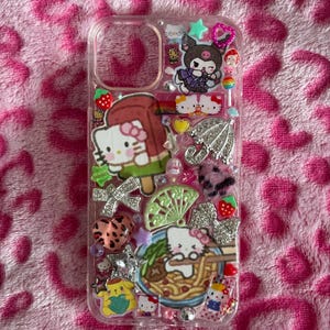 May include: Clear phone case decorated with various Hello Kitty and other cartoon character charms. The case features a Hello Kitty popsicle, a bowl of ramen, and a silver umbrella charm. Other charms include bows, stars, and strawberries.