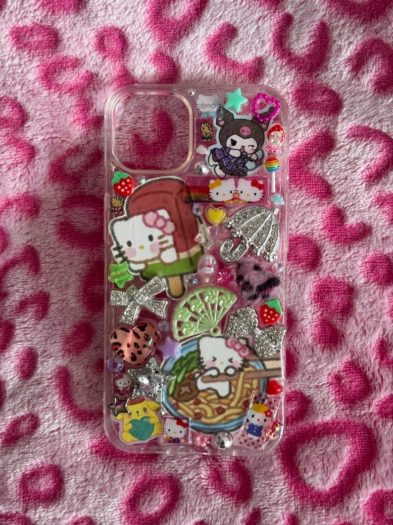 May include: Clear phone case decorated with various Hello Kitty and other cartoon character charms. The case features a Hello Kitty popsicle, a bowl of ramen, and a silver umbrella charm. Other charms include bows, stars, and strawberries.