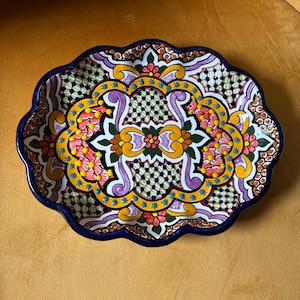 Puede incluir: Una fuente ovalada de cerámica con borde ondulado. La fuente presenta un vibrante diseño floral en tonos amarillos, rosas, morados y azules, sobre un fondo blanco. El borde es de color azul oscuro. Probablemente pintado a mano.