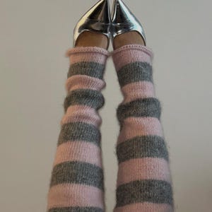 May include: Silver pointed-toe heels paired with pink and gray striped leg warmers. The leg warmers are knit with alternating bands of pink and gray. The shoes have a metallic finish.