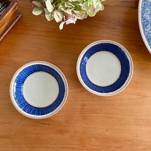 May include: Two small, round plates with a white centre and a blue, patterned border. The plates are on a wooden surface. The blue border has a textured design. The plates are likely for serving or display.