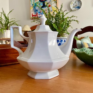May include: A white ceramic teapot featuring an octagonal body, a matching lid with a pointed finial, a curved handle, and a long spout. The base and the midsection of the teapot are decorated with a subtle beaded trim detail along the edges.