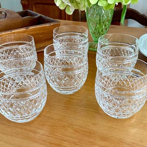 Puede incluir: Seis vasos de cristal tallado transparente con un patr&oacute;n de diamantes est&aacute;n dispuestos sobre una superficie de madera. Los vasos son redondos y de perfil bajo. Una bandeja de madera, un jarr&oacute;n de cristal verde y un plato blanco con fruta est&aacute;n en el fondo.