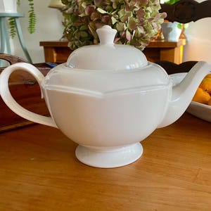 May include: A white ceramic teapot with a lid, spout, and handle. The teapot has a rounded body and sits on a small base. The teapot is on a wooden table.
