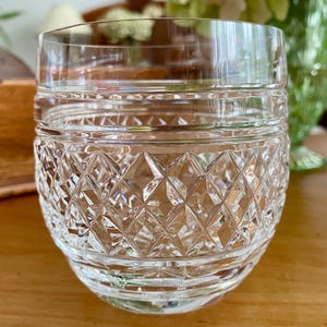 Puede incluir: Un vaso de cristal tallado transparente con un patr&oacute;n de diamantes en la mitad inferior y bandas horizontales cerca de la parte superior. El vaso est&aacute; sobre una superficie de madera, con un fondo borroso.