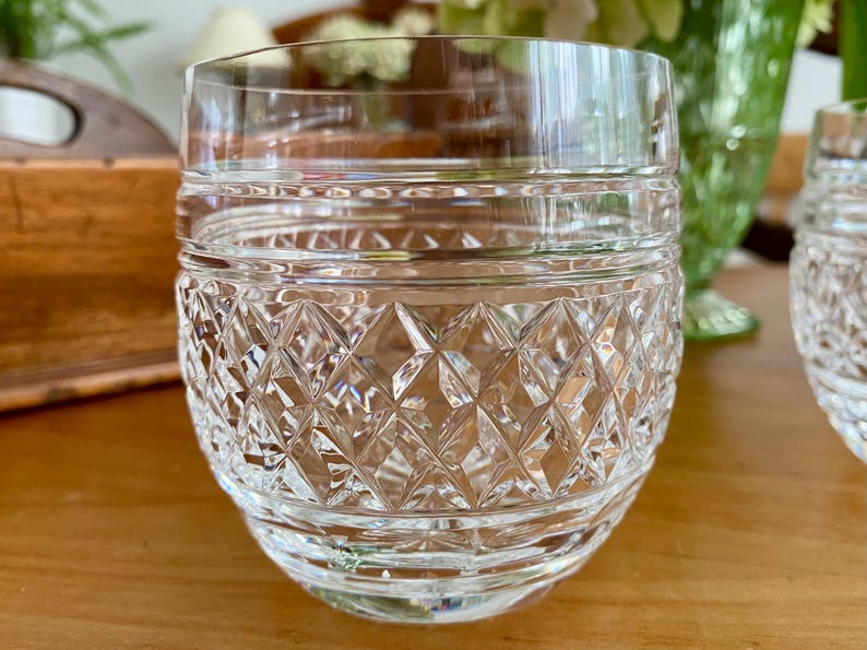 Puede incluir: Un vaso de cristal tallado transparente con un patr&oacute;n de diamantes en la mitad inferior y bandas horizontales cerca de la parte superior. El vaso est&aacute; sobre una superficie de madera, con un fondo borroso.