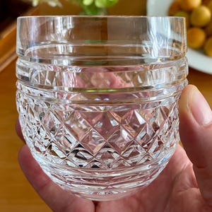 Puede incluir: Un vaso de cristal tallado transparente con un patr&oacute;n de diamantes y bandas horizontales. El vaso se sostiene en una mano, con un fondo borroso de fruta y vegetaci&oacute;n. Este vaso de estilo vintage es adecuado para servir bebidas.