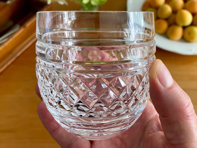 Puede incluir: Un vaso de cristal tallado transparente con un patr&oacute;n de diamantes y bandas horizontales. El vaso se sostiene en una mano, con un fondo borroso de fruta y vegetaci&oacute;n. Este vaso de estilo vintage es adecuado para servir bebidas.