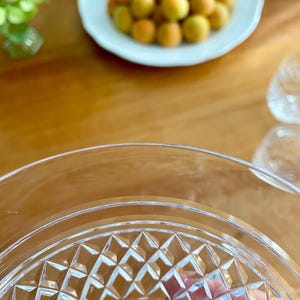 Puede incluir: Primer plano de un plato de cristal transparente con un patr&oacute;n de diamantes, con un fondo borroso de una mesa de madera, un plato blanco con albaricoques y peque&ntilde;os vasos transparentes. El plato tiene un borde decorativo.