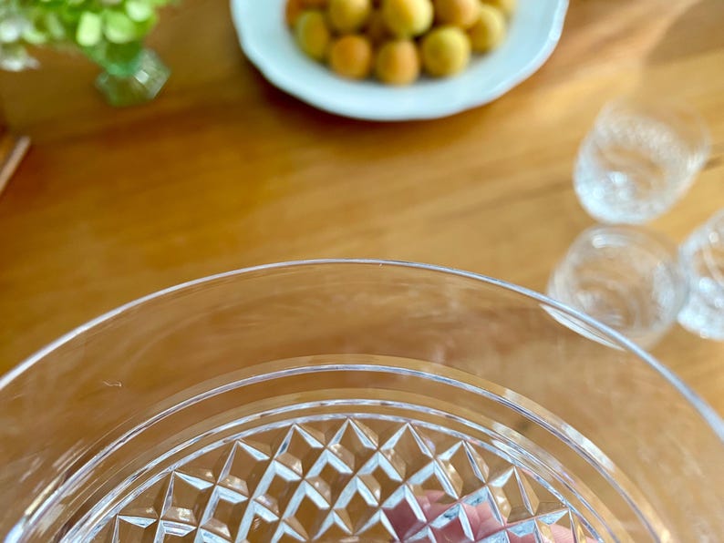 Puede incluir: Primer plano de un plato de cristal transparente con un patr&oacute;n de diamantes, con un fondo borroso de una mesa de madera, un plato blanco con albaricoques y peque&ntilde;os vasos transparentes. El plato tiene un borde decorativo.