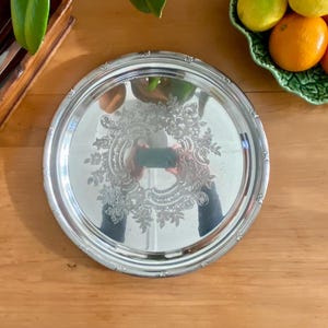 May include: A round silver-toned serving tray featuring a polished, reflective surface with an intricate, embossed floral and scrollwork pattern centred on the base and a decorative, beaded edge detail along the outer rim of the circular metal platter.