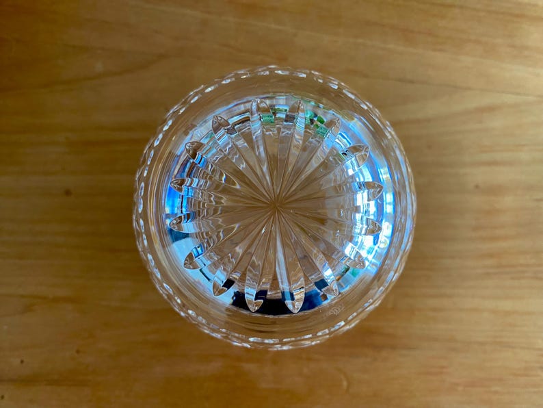 Puede incluir: Un vaso transparente y redondo con un patr&oacute;n grabado detallado. El vaso tiene un dise&ntilde;o de estrella en la parte inferior y un borde con cuentas. El vaso est&aacute; sobre una superficie de madera. Probablemente sea un vaso para beber o un objeto decorativo.
