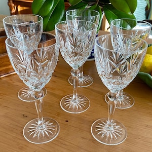May include: A set of five clear crystal wine glasses featuring a cut glass starburst pattern on the bowls and circular bases. Each glass has a slender stem and a faceted design that catches the light, creating a reflective and textured appearance.