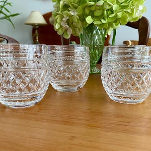 Puede incluir: Seis vasos de cristal tallado transparente con un patr&oacute;n de diamantes. Los vasos son cil&iacute;ndricos con una base redondeada. Est&aacute;n colocados sobre una mesa de madera. Un jarr&oacute;n de flores verdes est&aacute; en el fondo.