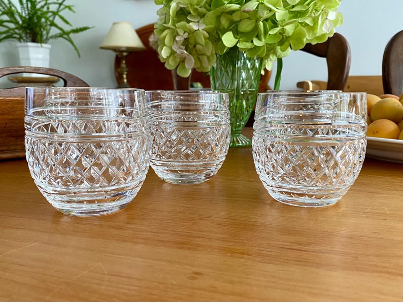 Puede incluir: Seis vasos de cristal tallado transparente con un patr&oacute;n de diamantes. Los vasos son cil&iacute;ndricos con una base redondeada. Est&aacute;n colocados sobre una mesa de madera. Un jarr&oacute;n de flores verdes est&aacute; en el fondo.