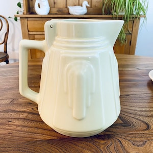 May include: A white ceramic jug with a fluted design. The jug has a handle and a spout. It is sitting on a wooden table.