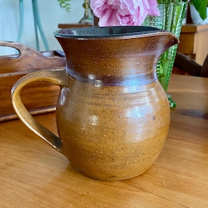 May include: A handcrafted, brown ceramic jug with a curved handle and a spout. The jug has a speckled texture and a dark brown rim. It sits on a wooden surface, with other decorative items in the background.