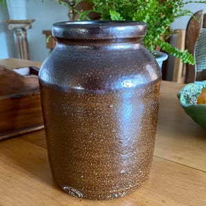 May include: A cylindrical stoneware crock featuring a two-tone glaze with a dark brown rim that transitions into a speckled, lighter brown body. The vessel has a wide, open mouth and a thick, sturdy base, showcasing a textured, earthy finish.