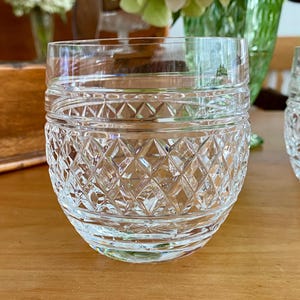 Puede incluir: Vaso de cristal transparente con un patr&oacute;n de corte de diamante. El vaso tiene una forma redondeada con una base gruesa y un borde liso. El vaso est&aacute; sobre una superficie de madera.
