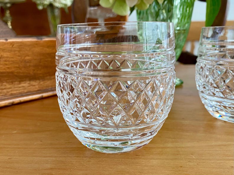 Puede incluir: Vaso de cristal transparente con un patr&oacute;n de corte de diamante. El vaso tiene una forma redondeada con una base gruesa y un borde liso. El vaso est&aacute; sobre una superficie de madera.